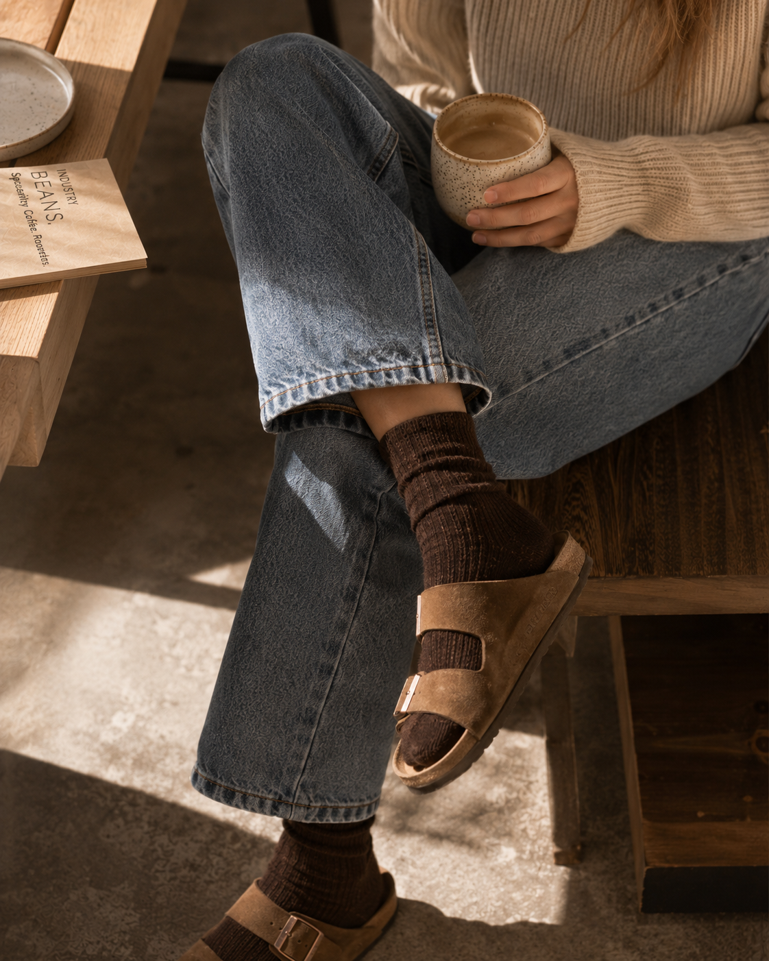 Person sitting on a wooden bench holding a cup, wearing jeans and sandals.
